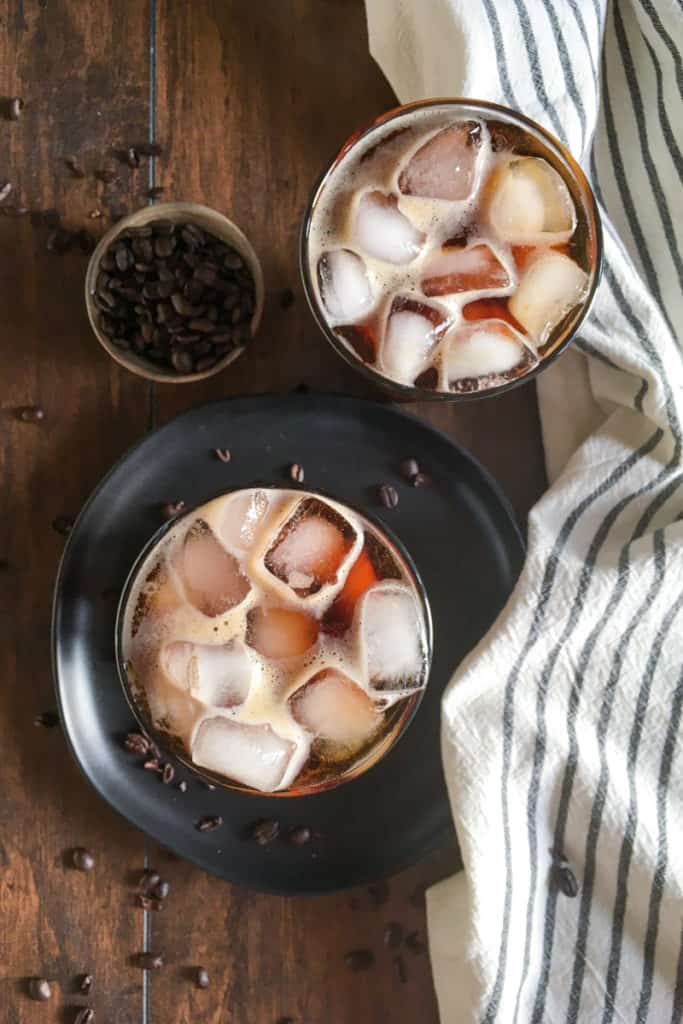 Overhead picture of two cups of cold brew coffee with ice floating on top and coffee beans scattered on table.
