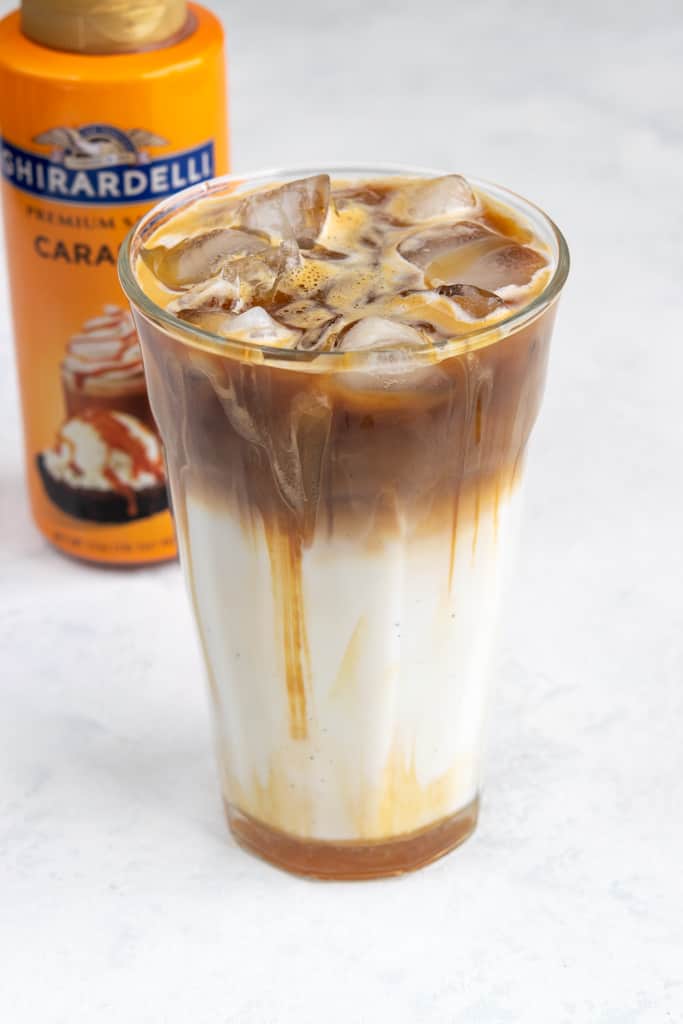 Bottle of ghirardelli sauce behind macchiato with caramel drizzled on top.