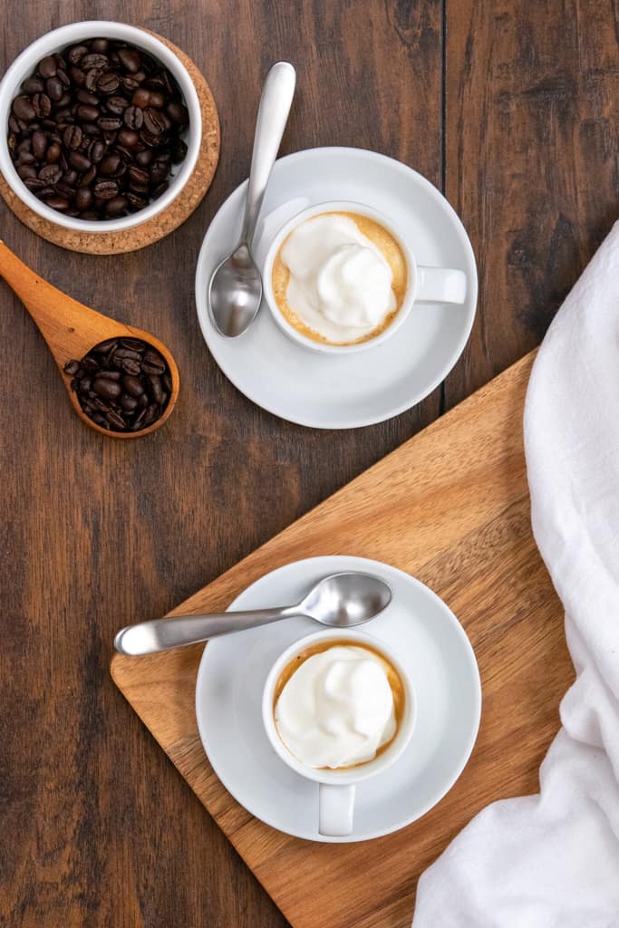 Overhead picture of two cups of espresso con panna and a scoop of coffee beans.