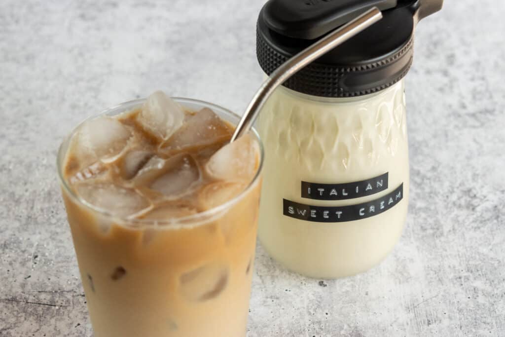 An iced coffee with cream and a straw in the cup in front of a jar of homemade coffee creamer that has an embossed label on it that says, Italian Sweet Cream.