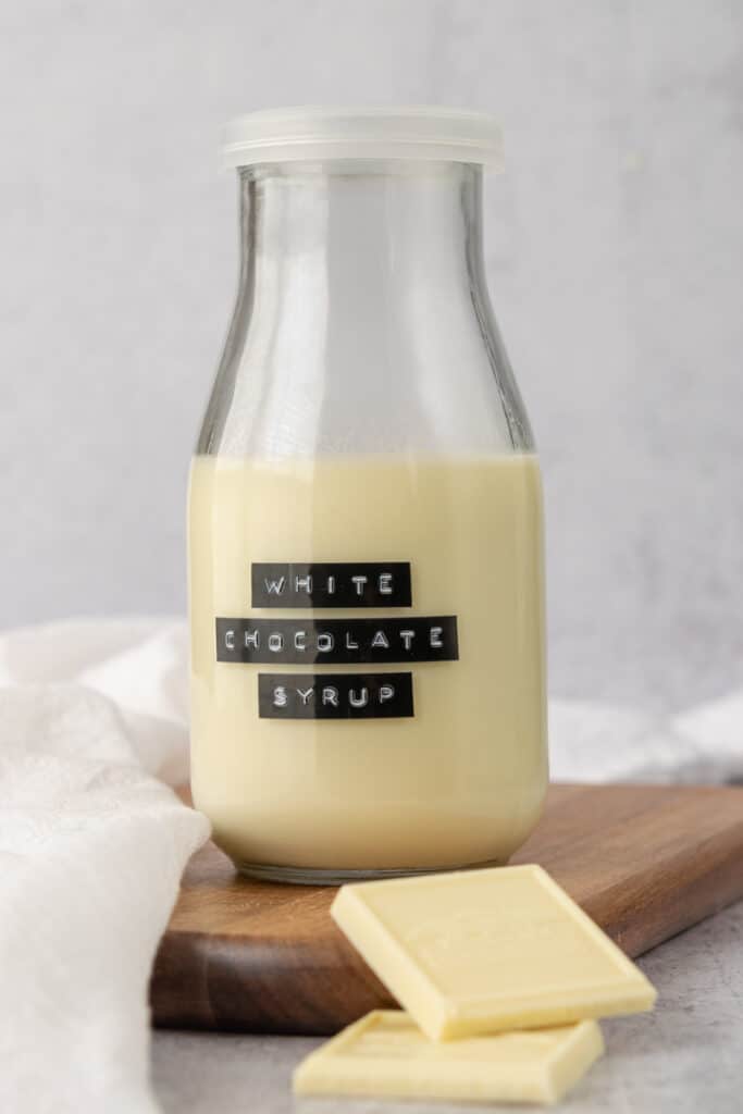 Homemade white chocolate syrup for coffee in a jar and two pieces of white chocolate bar.