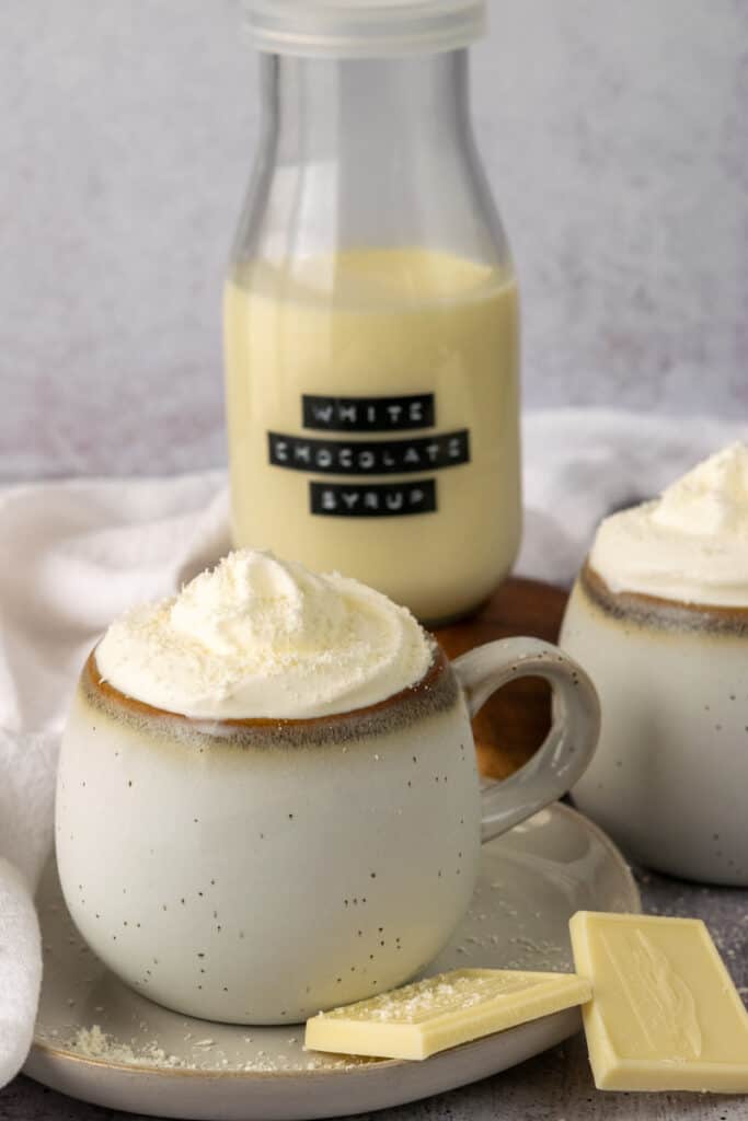 Two cups of whipped cream and white chocolate topped coffee sitting next to a jar full of white chocolate syrup.