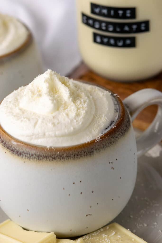 Cup of coffee topped with whipped cream and white chocolate shavings in front of a bottle of homemade white chocolate syrup for coffee.