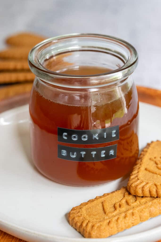 Open jar of cookie butter syrup sitting on a plate with two Lotus Biscoff cookies.