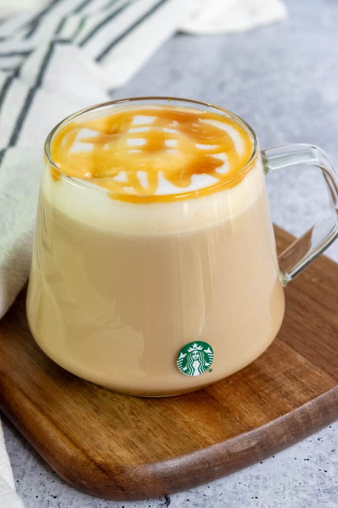 A hot Starbucks caramel macchiato made at home in a glass mug with a Starbucks logo, with a thick layer of caramel sauce on top.