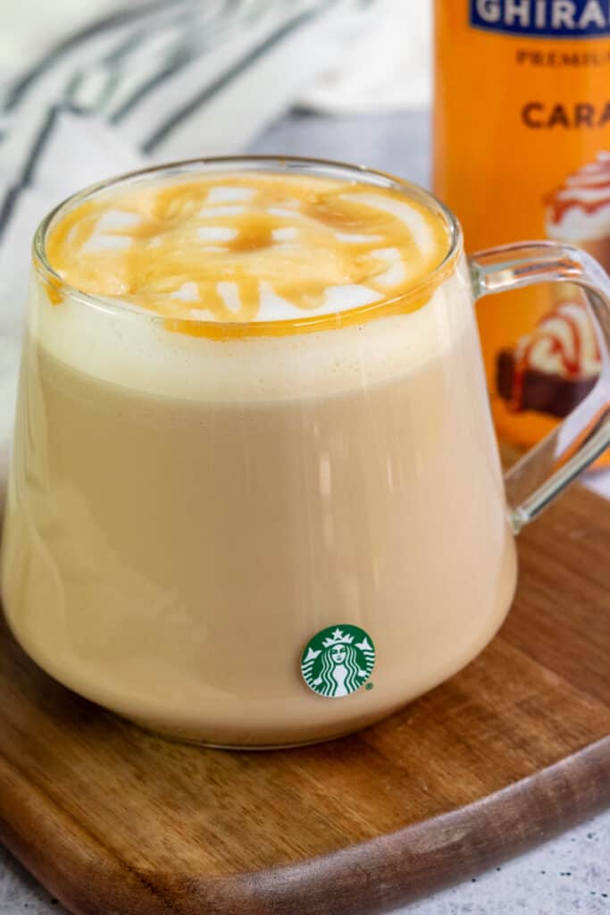A layered caramel macchiato with a crosshatch of caramel sauce on top and a bottle of Ghirardelli caramel sauce in the background.