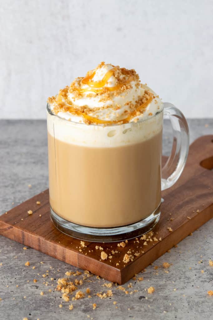 A decadent pumpkin pie latte in a glass mug layered with pumpkin spice syrup, coffee, milk, whipped cream, pie crust topping and caramel drizzle.