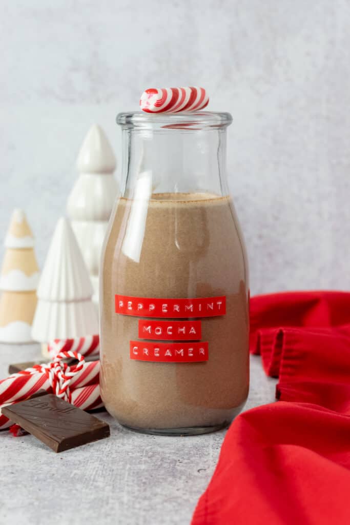 Peppermint Mocha Coffee Creamer in a glass bottle labeled, peppermint mocha creamer, sitting in front of decorative Christmas trees, peppermint candies and chocolate.