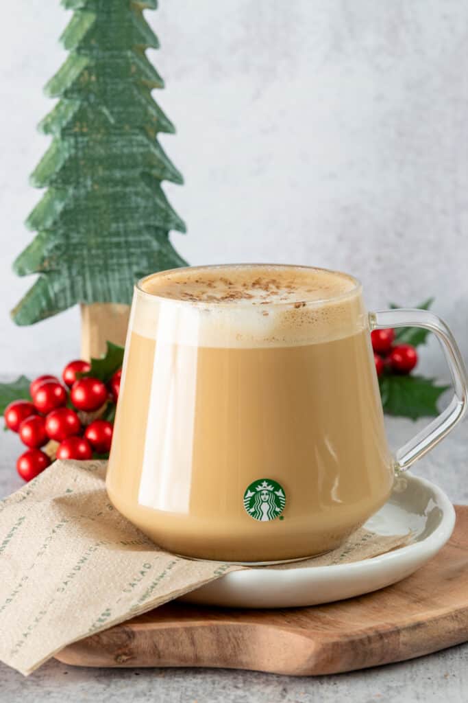 Eggnog latte in a glass Starbucks mug sitting on a Starbucks napkin with Christmas tree and holly decorations in the background.