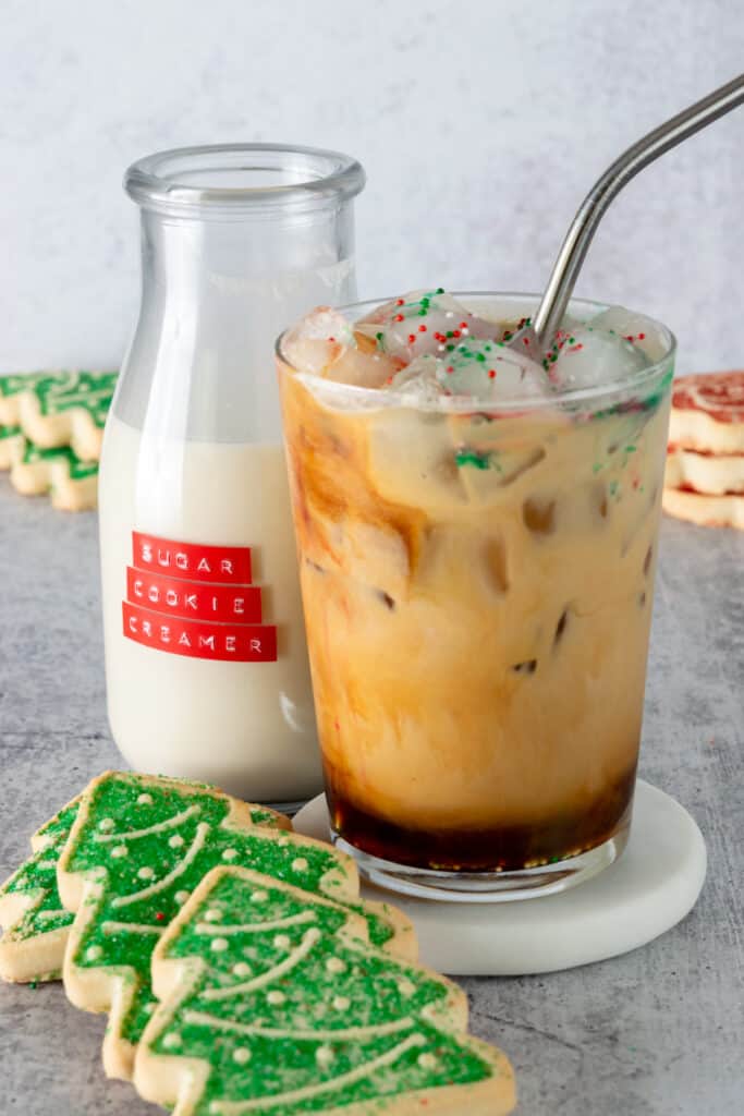 An iced sugar cookie latte next to a bottle of homemade sugar cookie coffee creamer and a Christmas tree-shaped sugar cookie.