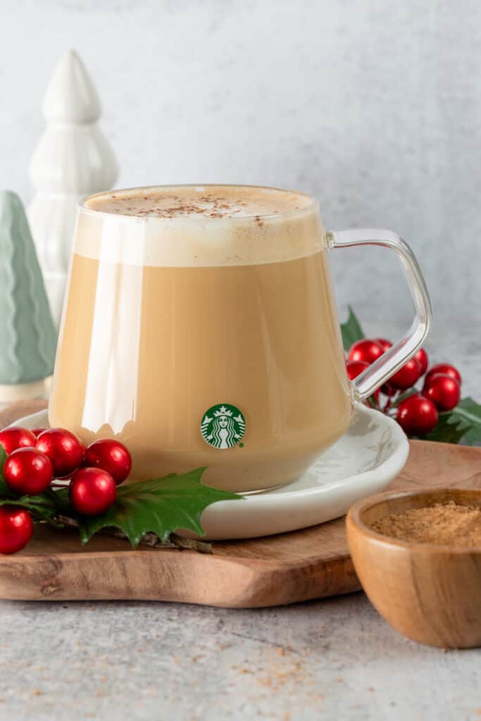 A homemade eggnog latte in a Starbucks cup with a small bowl of nutmeg next to it and Christmas tree decoration in the background.