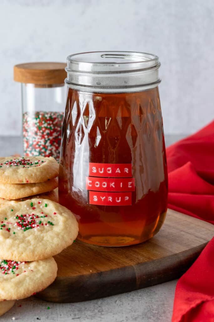 Starbucks sugar cookie syrup dupe in a glass jar with sugar cookie syrup label, surrounded by sugar cookies with sprinkles on top.