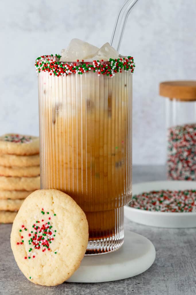 A festive iced sugar cookie latte in a glass whose rim has been dipped in sugar cookie syrup and red, green and white sprinkles.