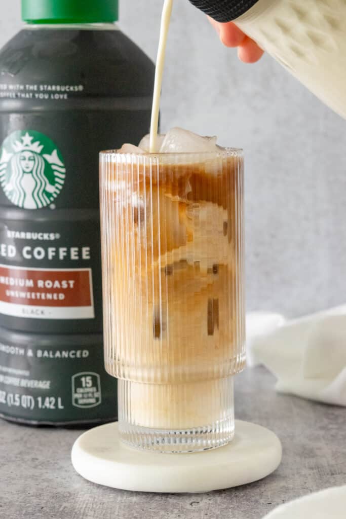 Vanilla sweet cream creamer being poured into a glass of iced coffee which is sitting in front of a bottle of ready-to-drink Starbucks iced coffee.