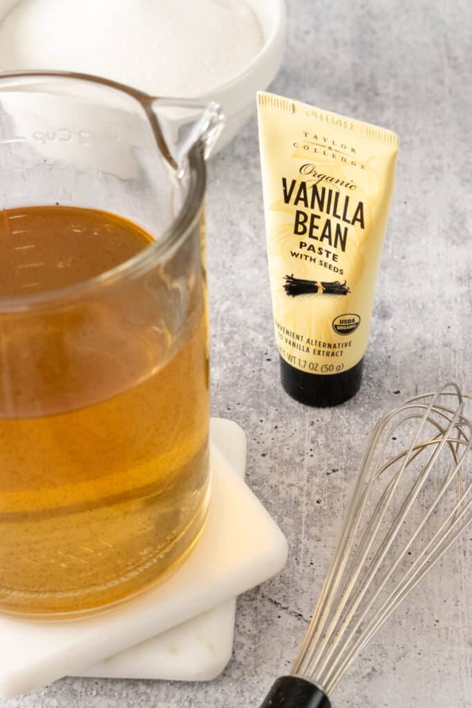 Vanilla bean paste, bowl of sugar, homemade vanilla syrup in a glass jar and a whisk.