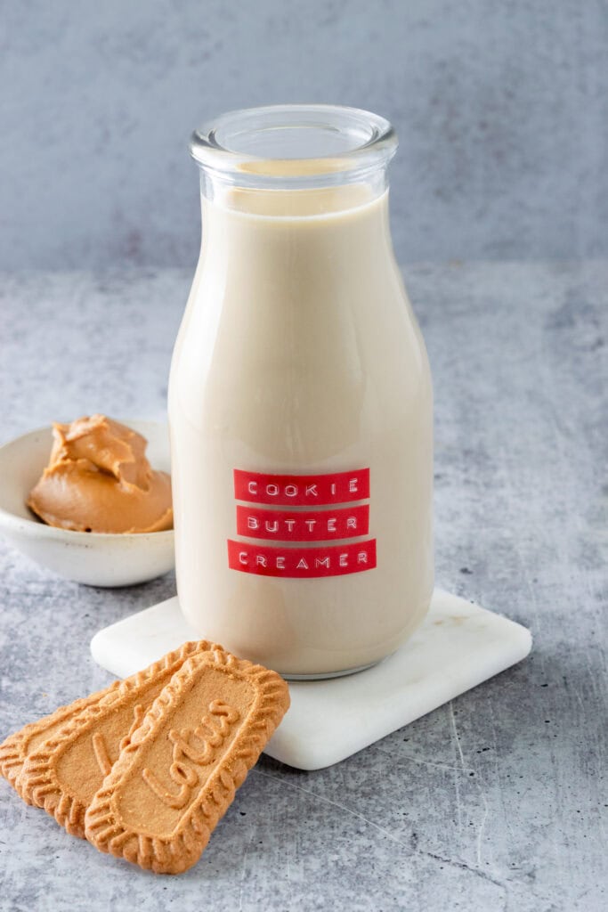 Bottle of homemade cookie butter coffee creamer next to 3 Lotus Biscoff cookies and a small bowl with cookie butter spread in it.